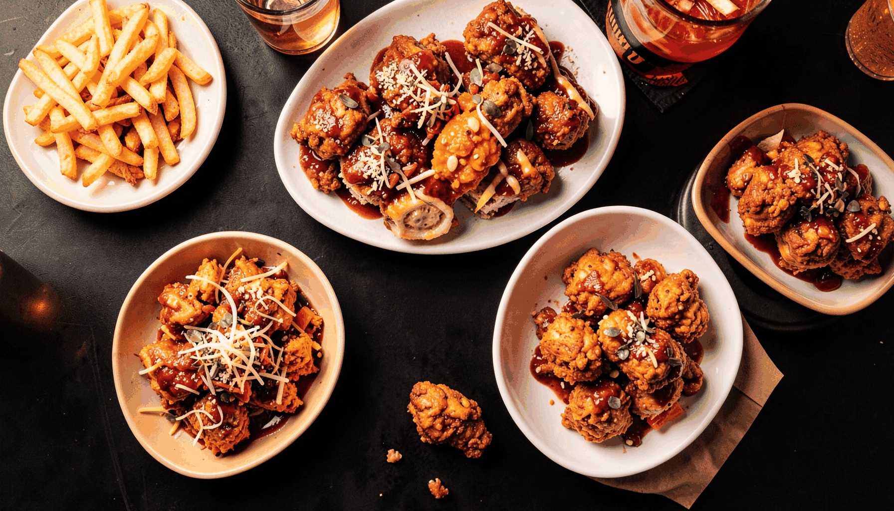 Overhead view of colorful comfort food dishes like loaded fries and saucy chicken bites arranged on a dark table, warm overhead lighting
